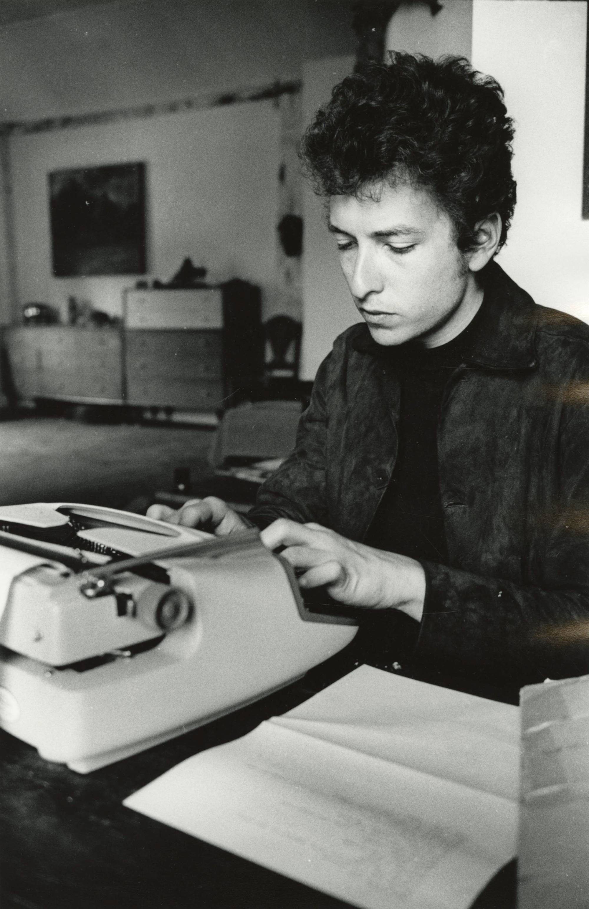 The photograph is in black and white. Bob Dylan sits at a desk typing at a typewriter.